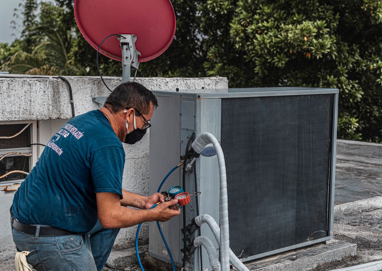 contact-img Technician repairing an air conditioner unit outdoors, wearing a facemask and using a manifold gauge.